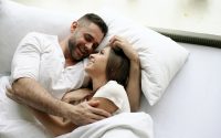 Young beautiful and loving couple kiss and hug into bed while waking up in the morning